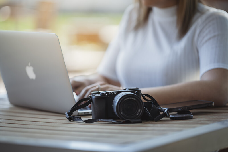Professional DSLR camera with strap on wooden desk next to open MacBook laptop.