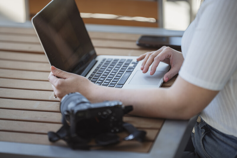 Photographer's hands using a laptop and wireless mouse at a wooden desk with a camera nearby.