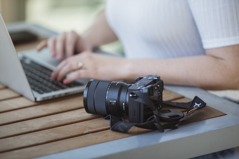 Professional camera with telephoto lens resting on wooden table beside laptop being used by photographer.