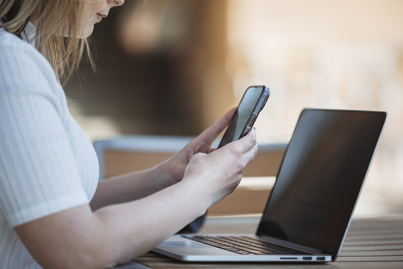 Woman holding a smartphone while working at a laptop outdoors in natural sunlight.