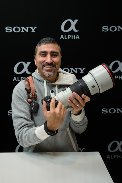 Photographer holding a white telephoto lens at a media event with Sony Alpha branding backdrop.