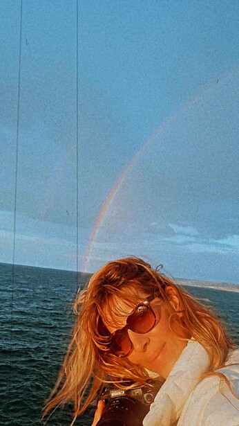 Woman wearing sunglasses on a boat with a rainbow visible over the ocean behind her.