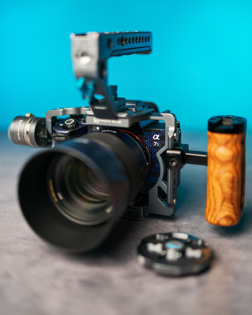 Professional cinema camera with large telephoto lens, mounted microphone, and wooden handle grip on gray surface.