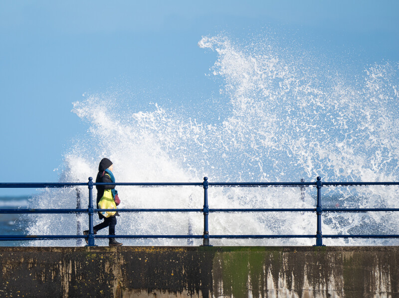 Person in yellow jacket standing at railing while large wave crashes against seawall.