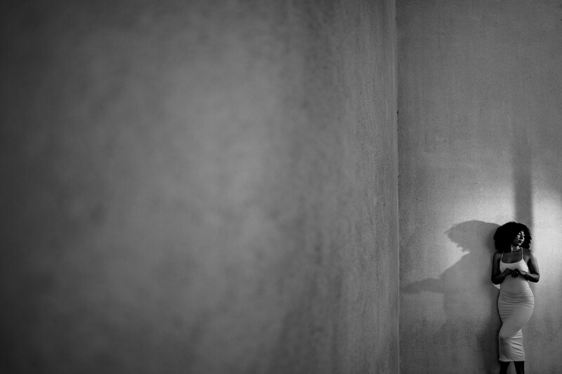 Woman in light dress standing against concrete wall casting dramatic shadow in black and white.