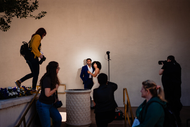 Behind-the-scenes of a professional photo shoot with crew members positioning lights and camera equipment around subjects in formal attire.
