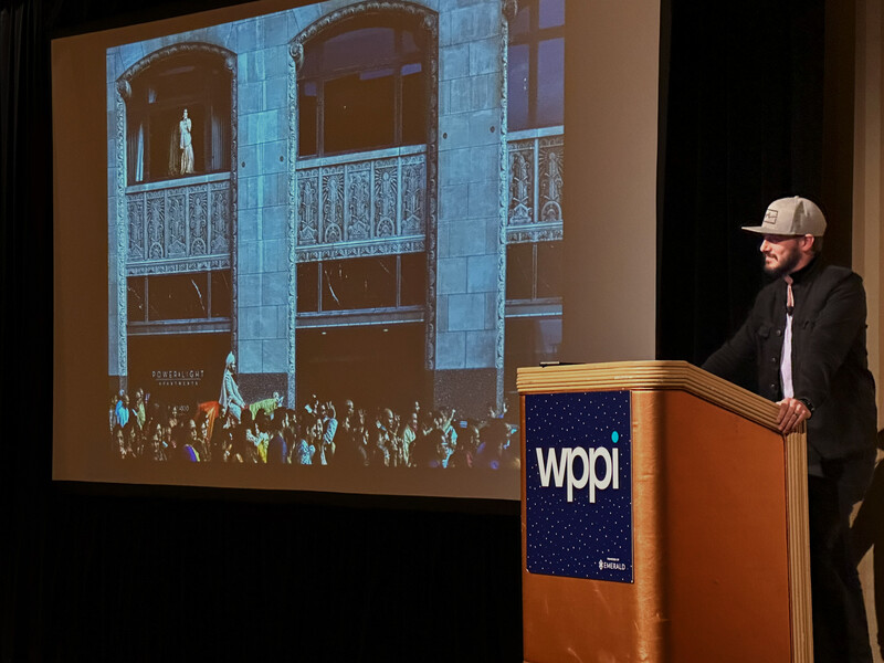 Speaker at wooden podium presenting architectural photograph projected on screen behind him.