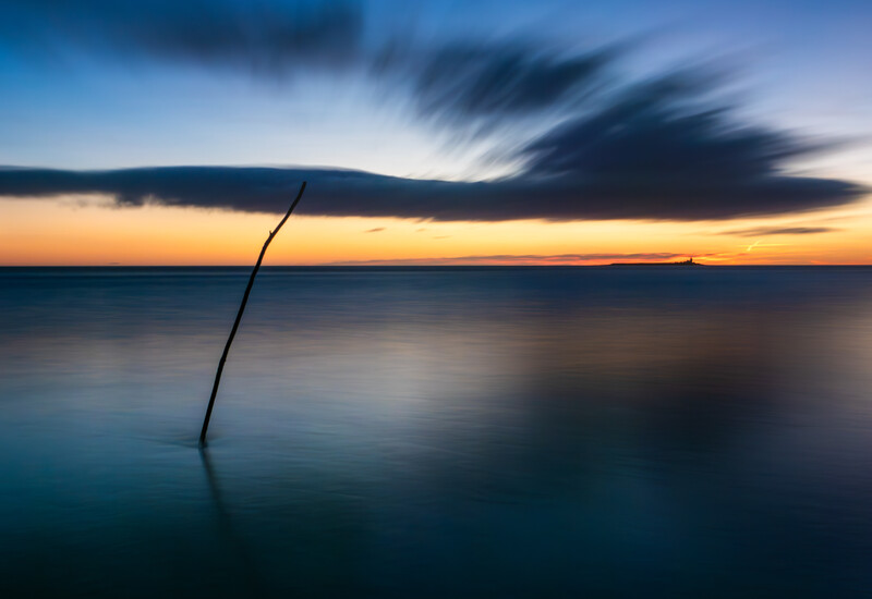 A solitary curved branch emerges from calm water during twilight, with dramatic clouds and a colorful sunset sky reflected below.