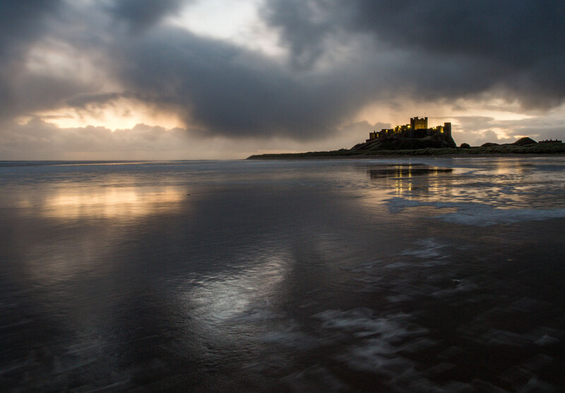 Medieval castle silhouetted on a rocky island under dramatic storm clouds at dusk.