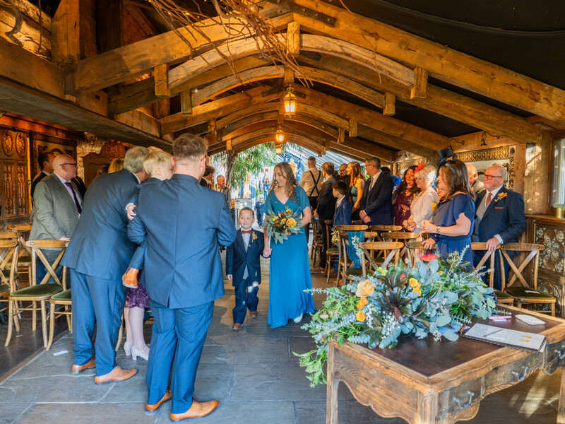 Wedding guests in formal blue attire gathered under wooden beams during an indoor reception ceremony.