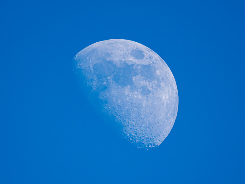 Waxing crescent moon against a deep blue daytime sky with visible lunar surface detail.