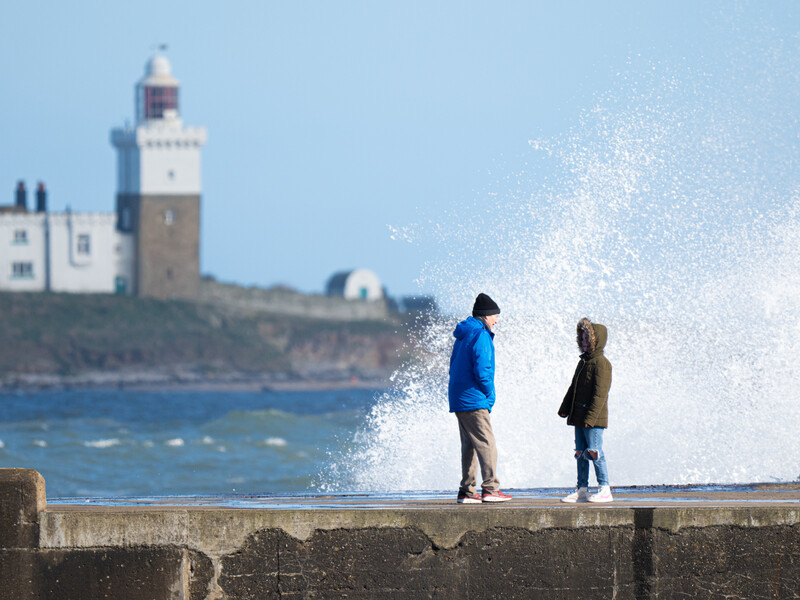 Two people standing on a seaside wall with a large wave splashing behind them and a lighthouse visible on a distant headland.