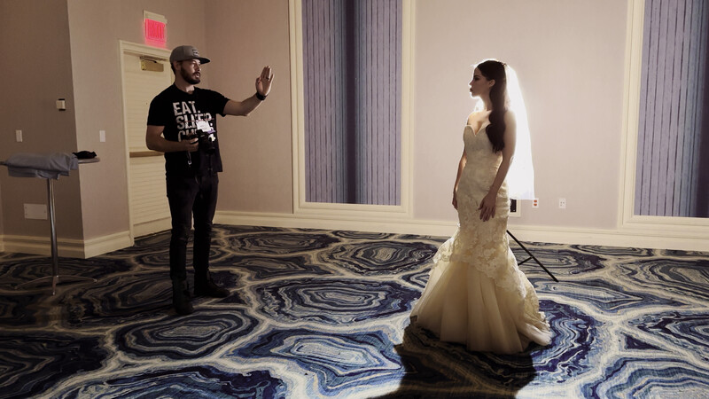 Man in black shirt gesturing while speaking to woman in white wedding dress in hotel ballroom.