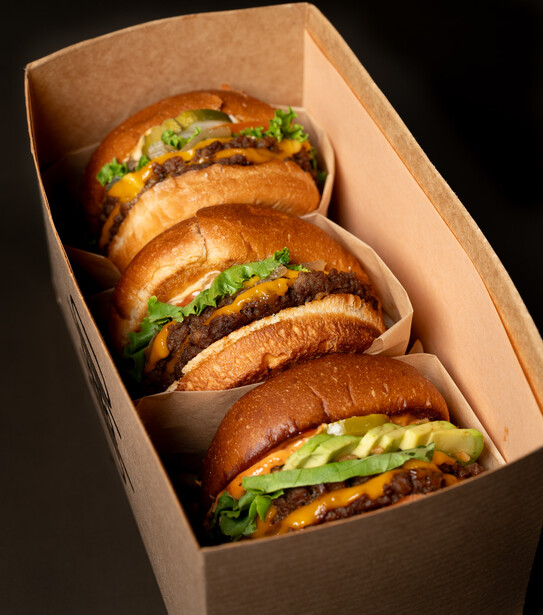Four gourmet sandwiches with melted cheese and fresh greens arranged in a cardboard takeout box.