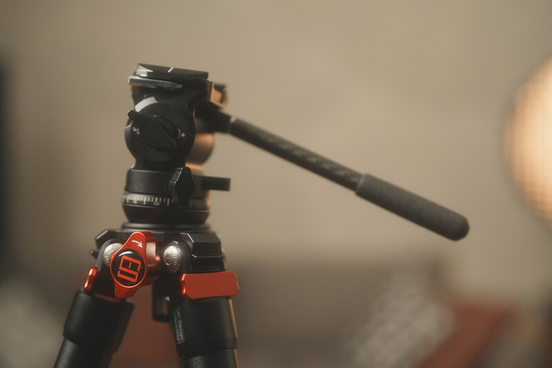 Professional video tripod with fluid head and red accents, shown in shallow focus with blurred background.