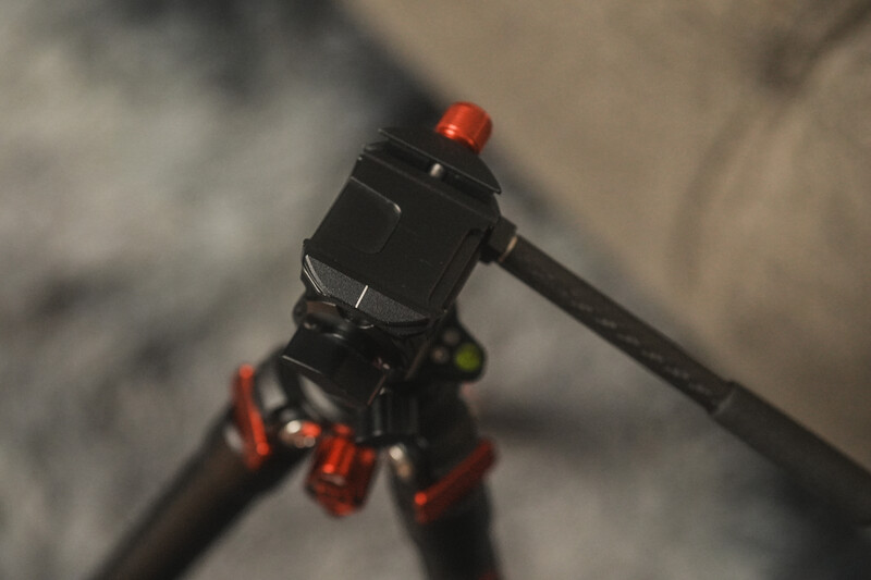 Close-up of a camera tripod head with red ball joints and adjustment knobs against a blurred concrete background.