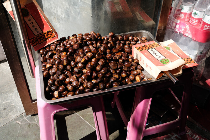Roasted chestnuts displayed in a metal tray on a pink vendor cart with an instructional sign nearby.