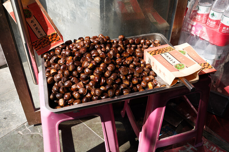 Large tray of roasted chestnuts displayed in a pink metal cart at an outdoor food stall.