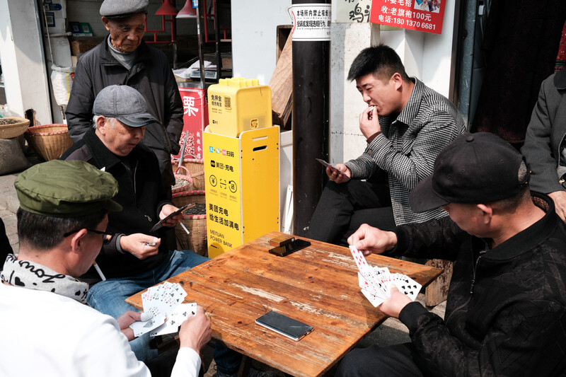 Five men gathered around a wooden table playing cards outdoors in an urban alley.