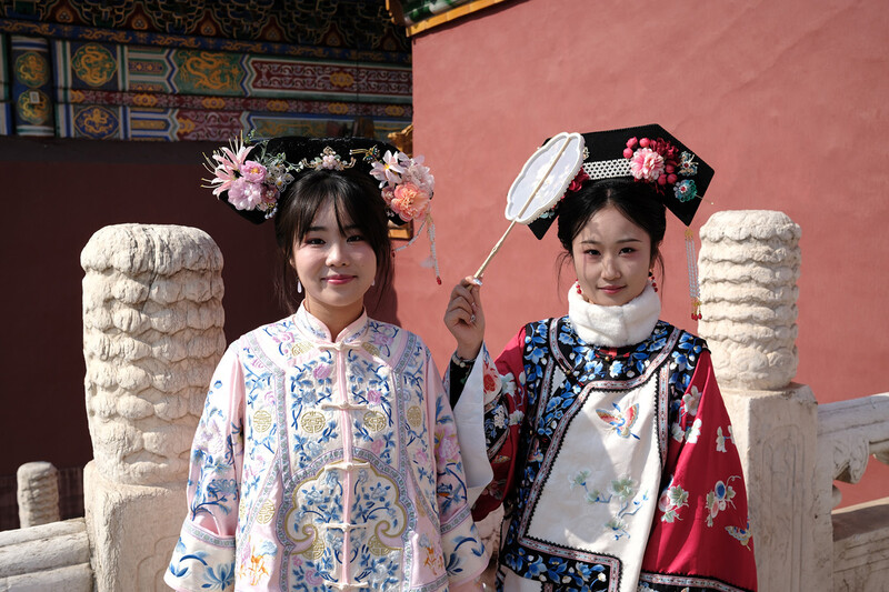 Two young girls in traditional embroidered garments and floral headdresses stand between ornate stone pillars against a pink wall.
