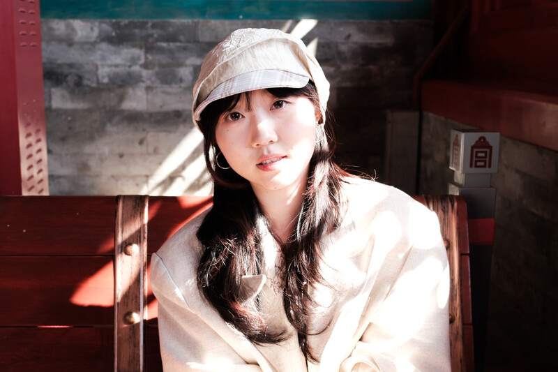 Young woman wearing a cream-colored cap and light jacket, photographed indoors with warm sunlight streaming across her face.