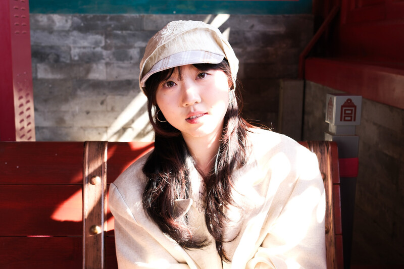 Portrait of a person wearing a cream-colored cap and jacket, lit by warm window light in an industrial interior.