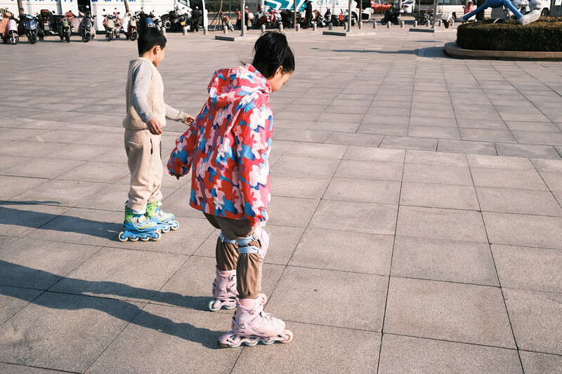Two young skateboarders standing on a wide plaza with long shadows cast across the pavement.