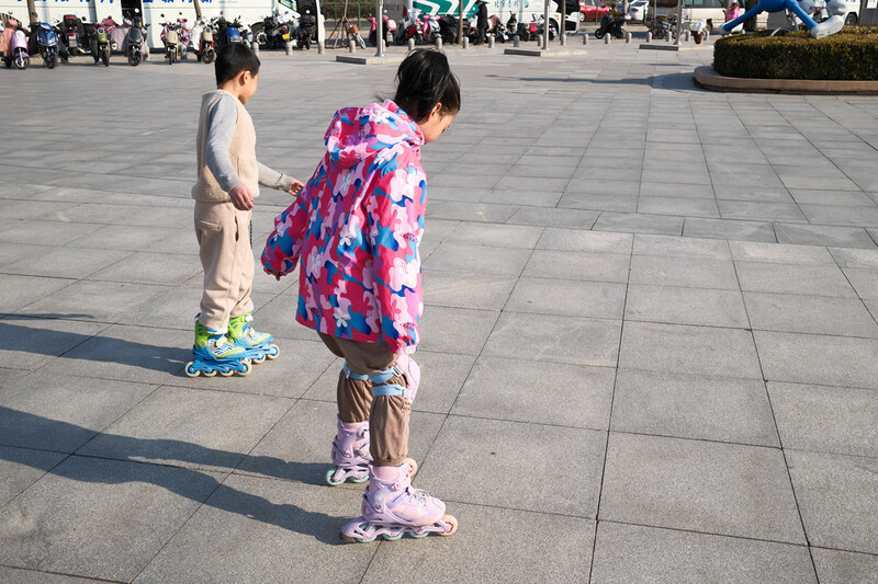 Two young children skateboarding in an open plaza on a sunny day.