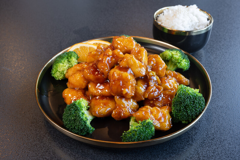Glazed chicken pieces arranged on a black plate with broccoli florets and a side of white rice.