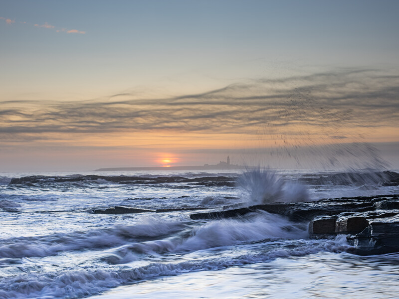 Ocean waves crash against rocky shoreline at sunrise, with dramatic spray and golden light reflecting off the water.