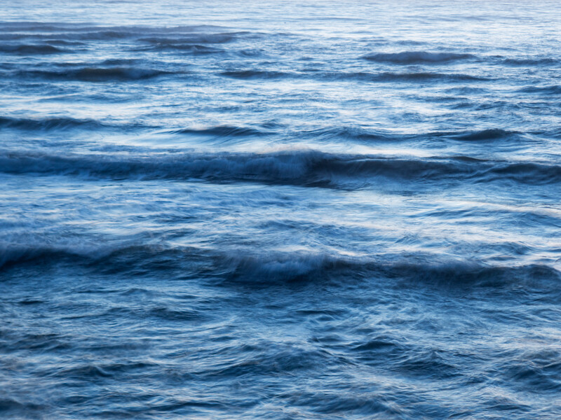 Ocean waves with multiple rows of white-capped swells rolling toward shore under overcast daylight.