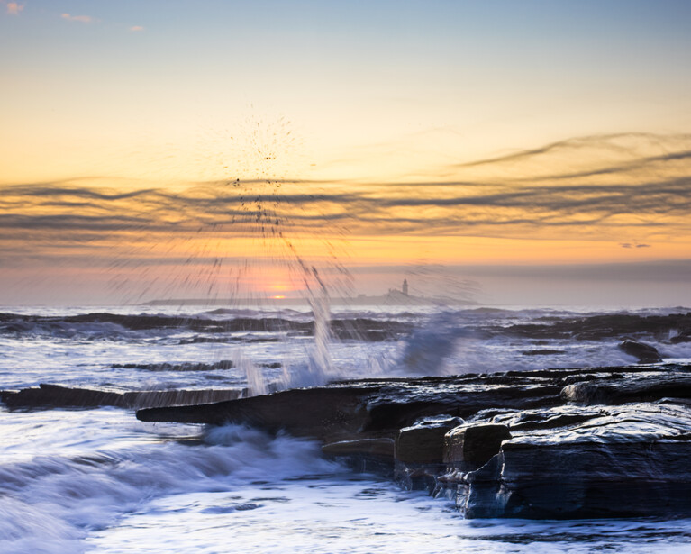 Ocean waves crashing against dark rocky shore at golden hour sunset.