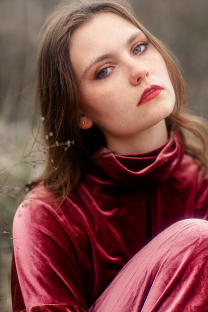 Young woman in burgundy velvet top with introspective gaze, outdoor setting.