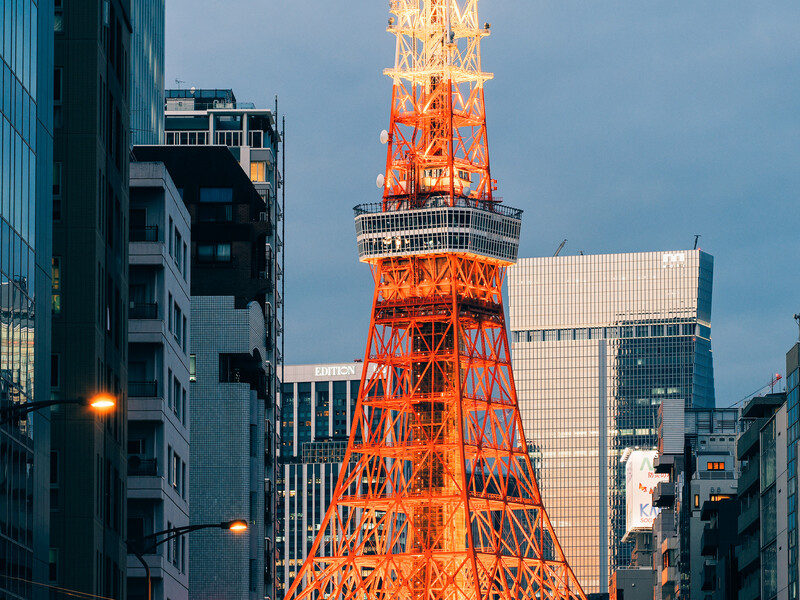 Illuminated red lattice tower rises among modern office buildings during twilight hour.