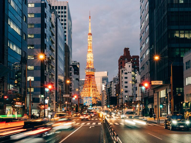 Tokyo street at dusk with illuminated tower centered between tall buildings and light trails from traffic.