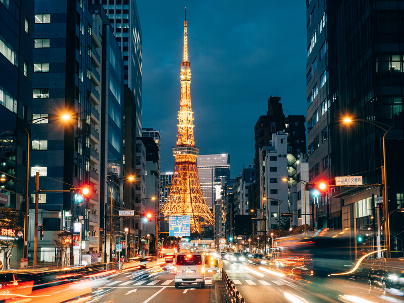 Illuminated red and white tower framed by tall buildings at twilight with light trails from traffic.