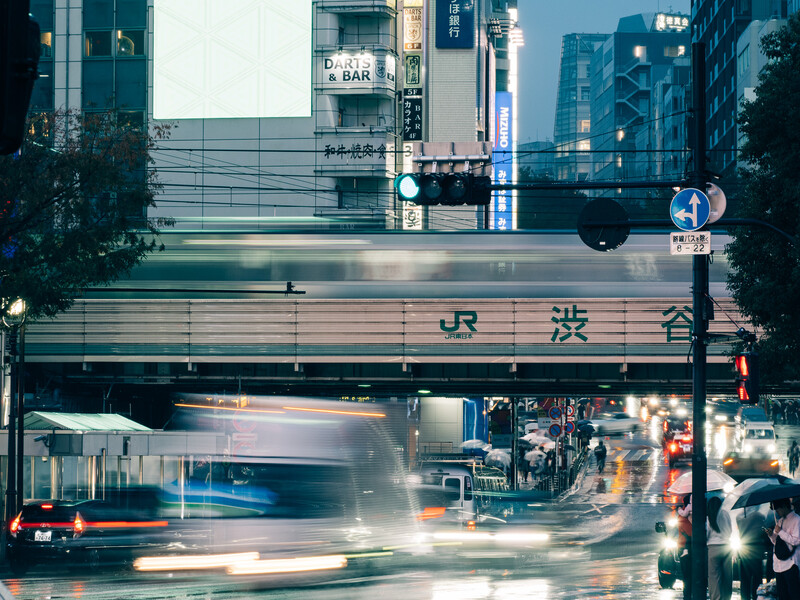 Long exposure photograph of a train station overpass with motion-blurred vehicles and pedestrians below in an urban street scene.