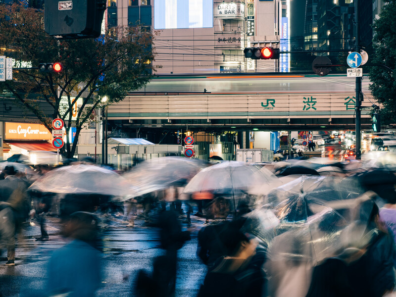 Blurred pedestrians crossing a wet street in front of a JR train station building in an urban area.