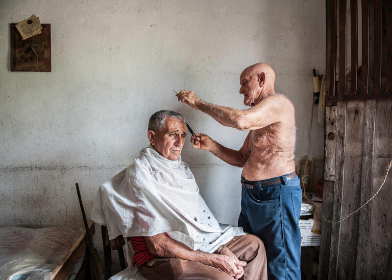 Older man seated getting a haircut from a standing barber in a sparse room.