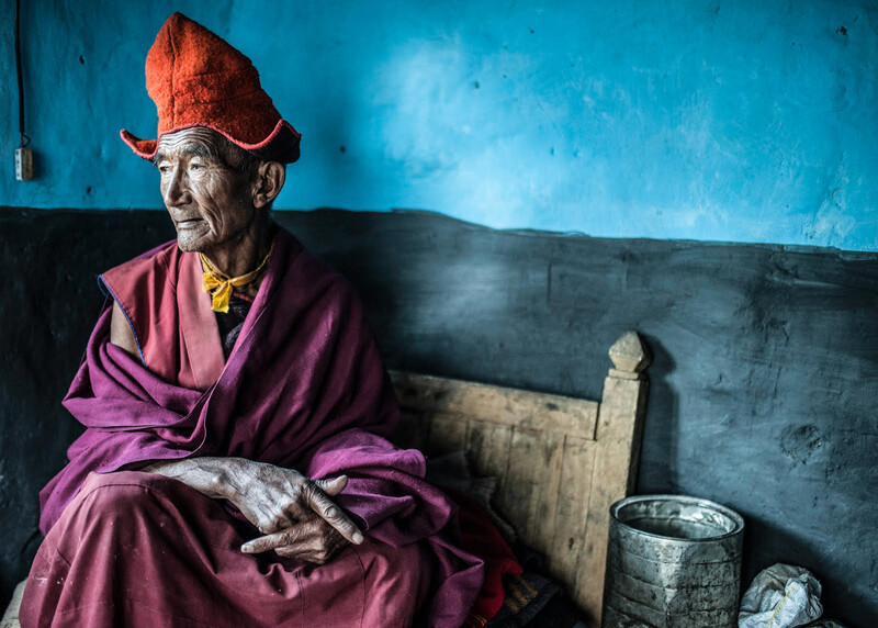 Elderly monk in burgundy robes and red hat seated against a blue wall, gazing thoughtfully to the side.