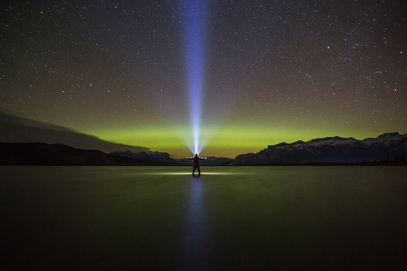 Lighthouse beam piercing night sky above calm water, with aurora borealis and stars visible overhead.