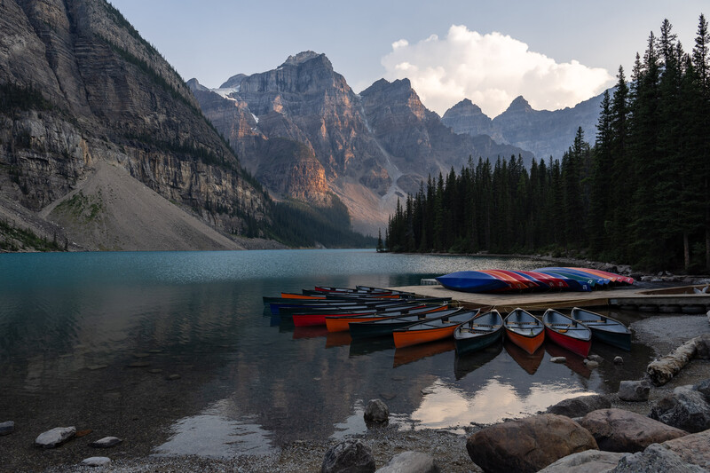 Colorful rental canoes lined up on the shore of a turquoise alpine lake surrounded by evergreen forests and dramatic mountain peaks.