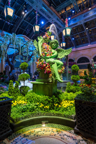 Large topiary sculpture illuminated with green lighting in an indoor atrium surrounded by holiday decorations.