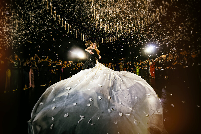 Bride in voluminous ball gown with pearl embellishments stands center stage beneath ornate ceiling with spotlights and crowd surrounding.