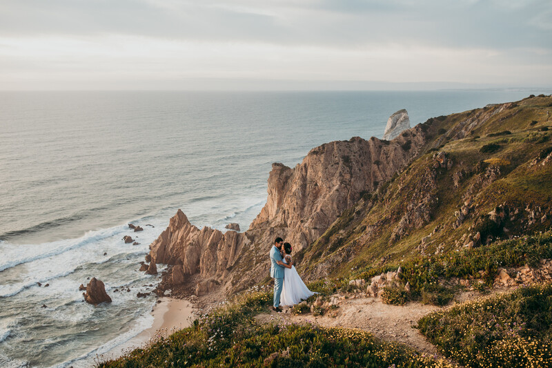 Couple in wedding attire standing on a clifftop overlooking rocky coastline and ocean.