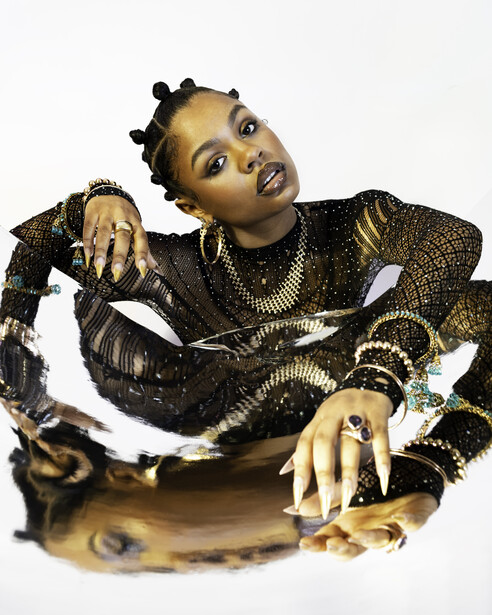 Person wearing ornate black and gold outfit with dramatic makeup and styled hair against white background.