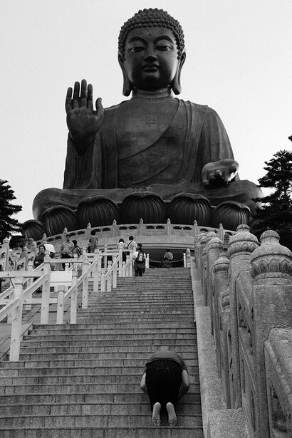 Large stone Buddha statue with raised hand blessing gesture, viewed from below with stairs leading upward in black and white photography.