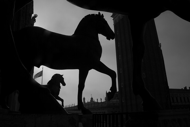 Silhouetted horse statue in architectural foreground with cityscape and smaller horse sculpture visible through arched openings.