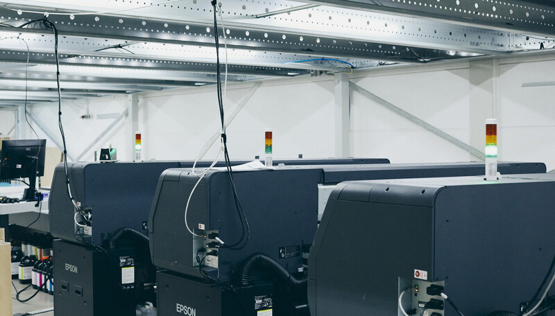 Multiple large-format industrial printers in a factory setting with exposed metal ceiling infrastructure.