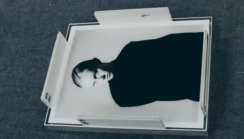 Black and white portrait of a person in dark clothing against white background, shown inside an open white box or frame.
